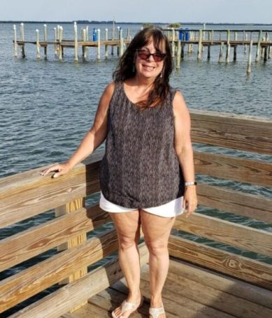 Photo of Pamela Green smiling on a wooden pier by the sea on a sunny day, wearing sunglasses and casual attire, with docks and calm water in the background, reflecting trust and confidence in growth mindset coaching.